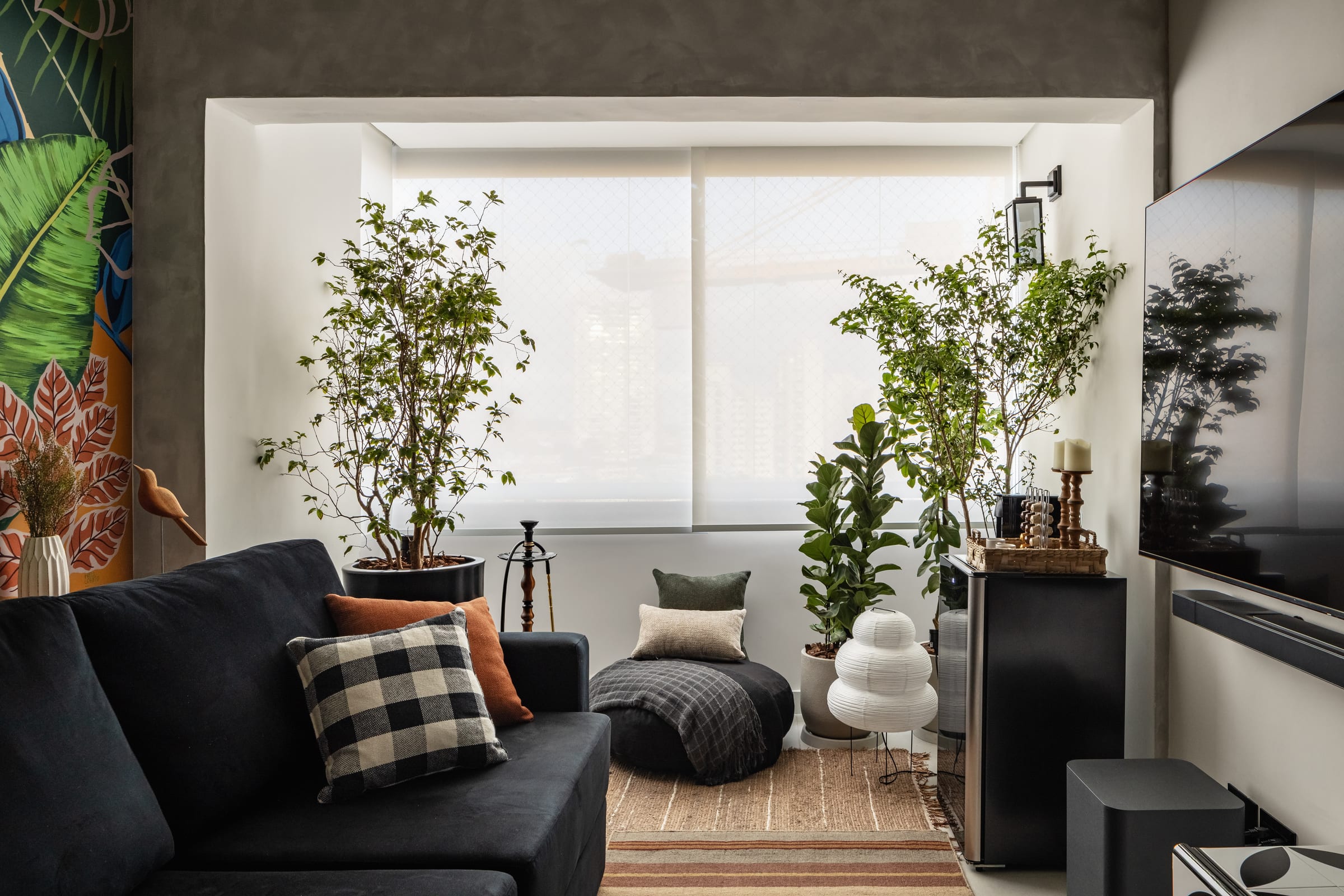 Estar de Erick e Ben com a varanda integrada tomada por plantas e árvores, sofá escuro e mural tropical, projeto INSIDE